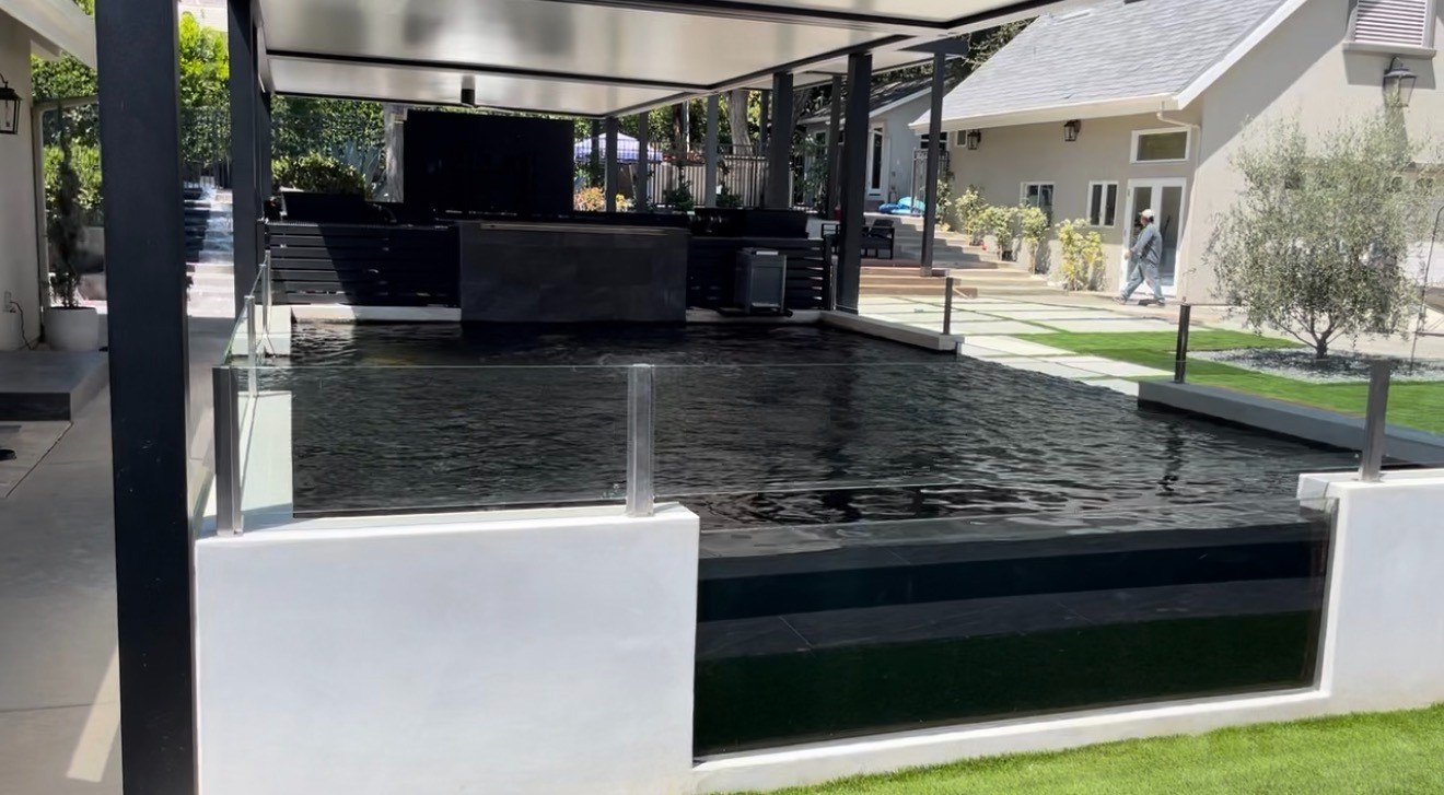 A backyard pool with a clear acrylic viewing panel set into the white retaining wall, the water visible through it from the lawn below.