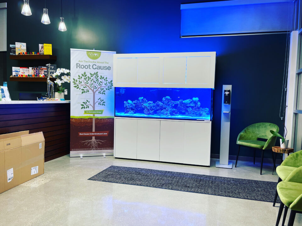 A blue-lit aquarium with rock formations on a tall white cabinet in a reception area with green chairs.